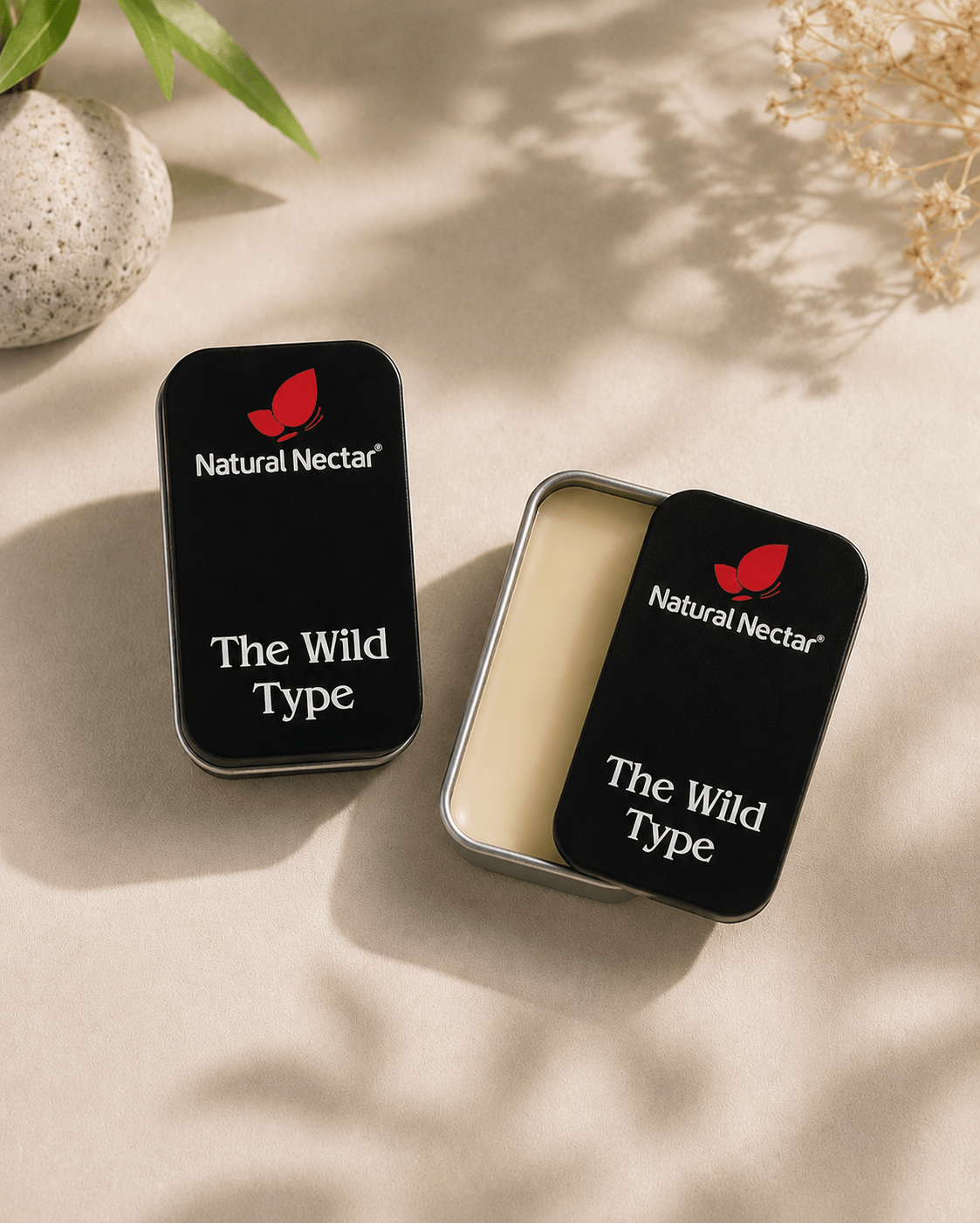 Two small rectangular metal tins with black lids labeled Natural Nectar 'The Wild Type' are arranged on a warm beige surface with soft leaf shadows. One tin is closed and the other is open, showing a pale cream-colored product inside.