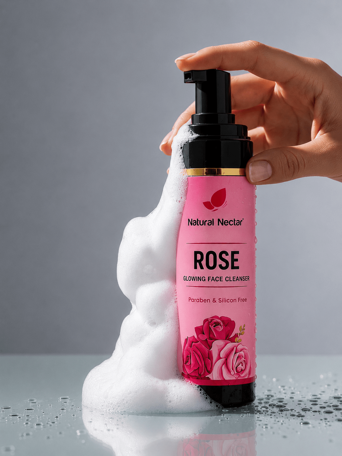 A hand holding a bottle of Natural Nectar Rose Glowing Face Cleanser with white foam lather flowing down the side, against a grey background.