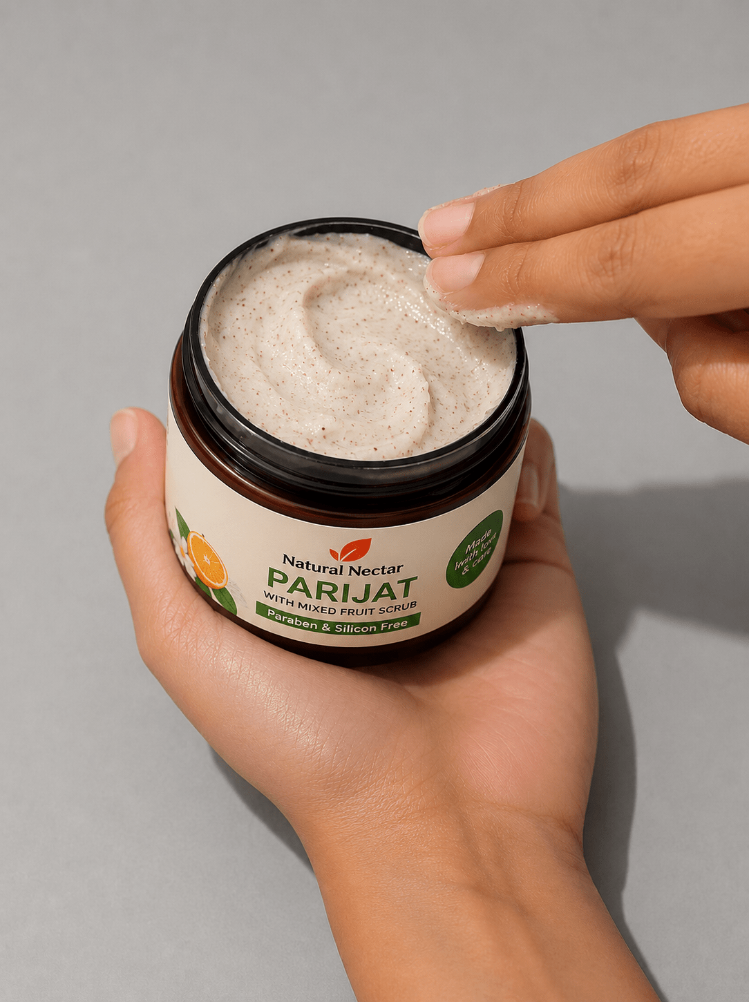 A hand holds an open jar of Natural Nectar Parijat Scrub showing the creamy beige scrub texture being applied.