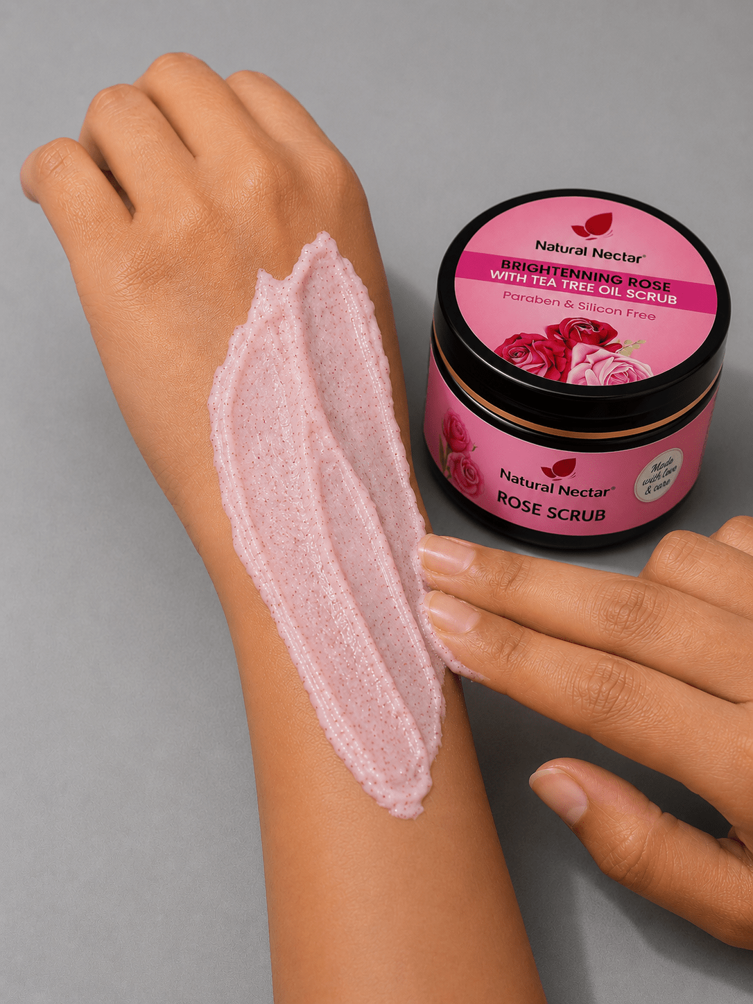 Close-up of a person's forearm with a thick swipe of pale pink rose scrub applied, next to pink jar on grey background.