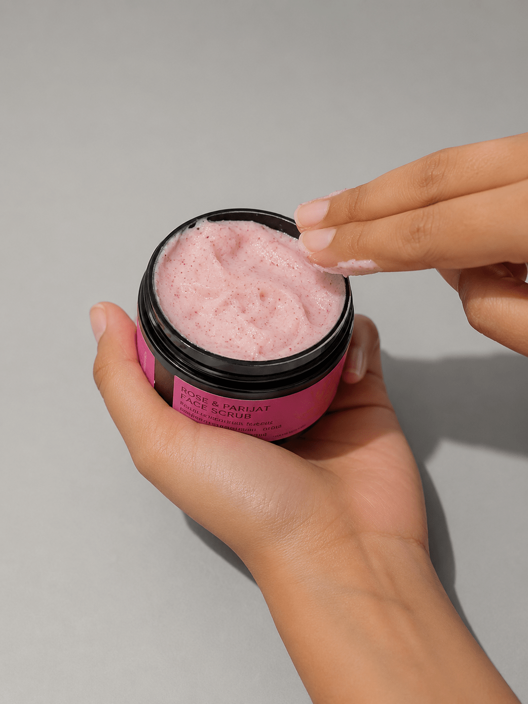 A hand holds an open jar of pink facial mask cream showing the textured cream, with another hand applying it.